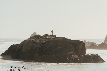 Person Standing on Rock Formation By The Seashore