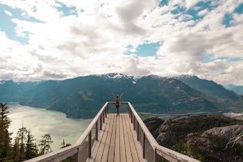 Person Standing With Hands on Air on Brown Wooden Dock With Overlooking View of Lake Under White Clouds and Blue Sky