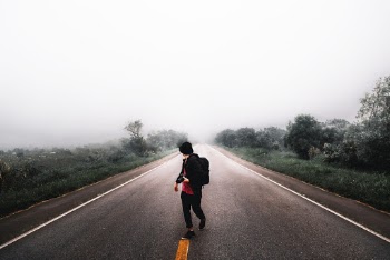 Person Waling in the Middle of Road in Between Grass Field Under White Skies
