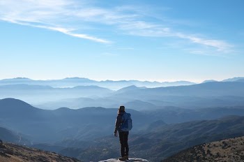 person wearing backpack standing on cliff in front of mountains during daytime