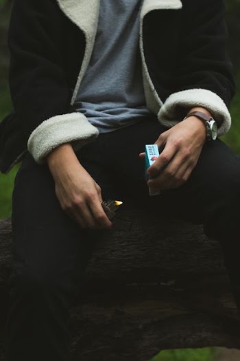 person wearing black and white jacket and black pants holding flip lighter and cigarette pack while sitting on tree log