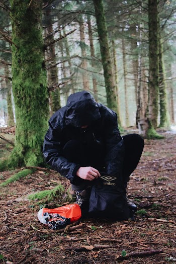 Person Wearing Black Hooded Jacket Sitting While Holding Black Umbro Drawstring Bag Surrounded With Green Trees
