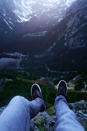 Person Wearing Denim Pants and Black Sneakers Sitting on Rock Cliff Viewing Mountains