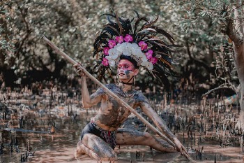 Person Wearing Feather Headdress Holding Wooden Stick While Kneeling on Mud