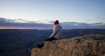 Person Wearing Gray Hooded Jacket and Black Pants Sitting on Mountain Cliff during Sunset