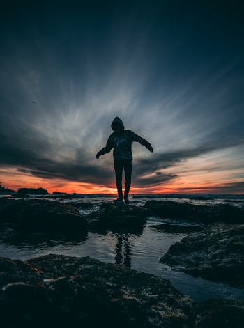 Person Wearing Hoodie Standing on Rock Surrounded by Body of Water