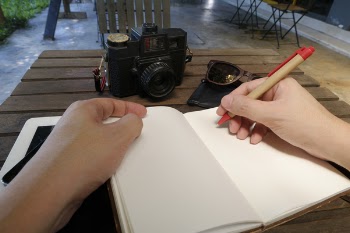 Person Writing Beside Dslr Camera on Brown Wooden Table
