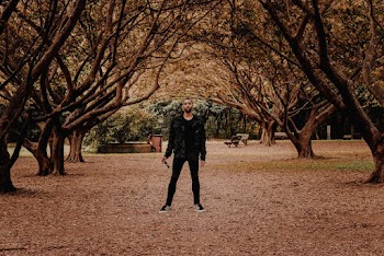 Photo of a Man Holding Sunglasses Standing Under Trees in a Park