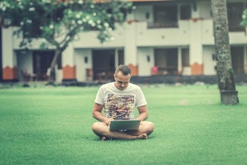Photo of a Man in White Printed T-shirt  Sitting on Green Grass in view  of a Building Using a Laptop 