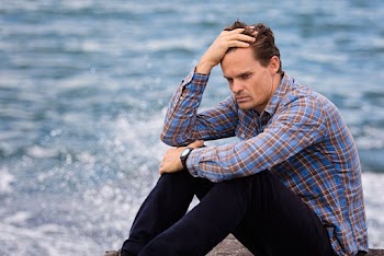 Photo of a man sitting near a body of water holding his head