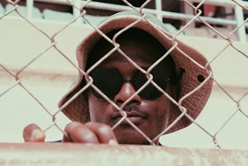 Photo of a man wearing black sunglasses standing behind a chain link fence