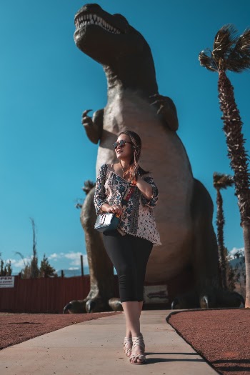 Photo of a woman wearing a printed top and sunglasses standing  near dinosaur figurine