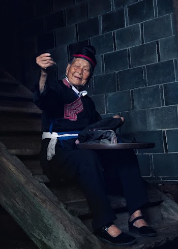 Photo of an elderly woman sitting on wooden stairs  sewing a cloth
