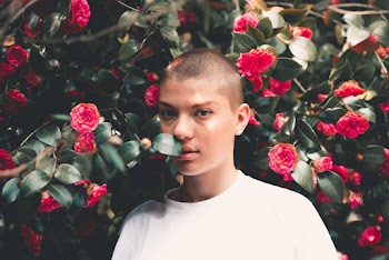 Photo of Bald Woman in White T-shirt Standing Beside Red Flowers