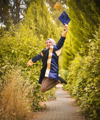 Photo of Happy Woman in Blue Academic Regalia and Hijab Jumping While Holding Her Graduation Cap by Footpath In Between Green-leafed Hedges