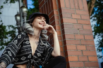 Photo of Laughing Woman in Black Top Posing with Her Eyes Closed Next to Brick Pillar