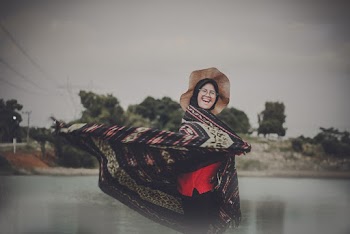 Photo of Laughing Woman in Hat and Hijab Standing by Body of Water While Flipping Her Shawl
