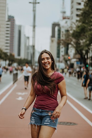 Photo of Laughing Woman in Maroon T-shirt and Blue Denim Shorts Standing on Sidewalk
