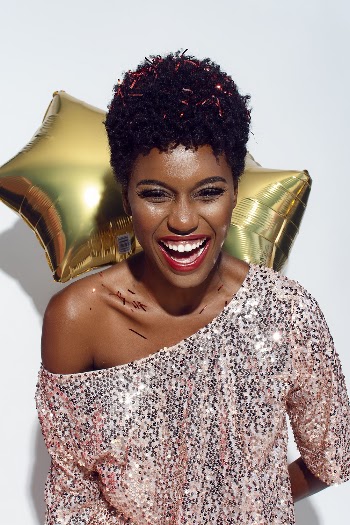 Photo of Laughing Woman with Confetti on Her Hair and Shoulders Posing In Front of White Background While Holding Golden Star Balloons Behind Her Back