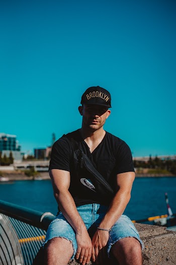 Photo of Man in Black Cap, T-shirt, and Blue Denim Shorts Sitting on Concrete Surface Posing with Body of Water in the Background