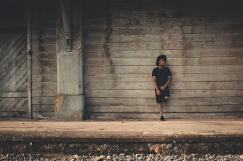 Photo of Man in Black T-shirt, Shorts, and Fedora Leaning on Wooden Wall Posing While Looking Away