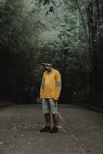 Photo of Man in Bunny Mask, Yellow Sweatshirt, and Cargo Shorts Standing Alone in the Middle of Empty Road In Between Trees