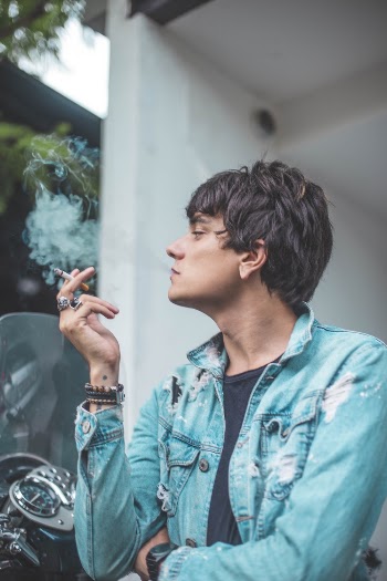 Photo of Man in Distressed Blue Denim Button-up Jacket Looking Away While Smoking a Cigarette