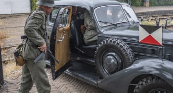 Photo of Man in Old Military Uniform Carrying a Rifle Standing By Vintage Military Car With Soldiers Inside