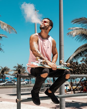 Photo of Man in Pink Sleeveless T-shirt and Black Jeans Holding Skateboard and Sitting on Metal Railing While Blowing Smoke