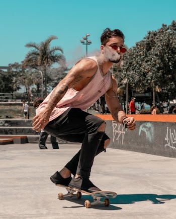 Photo of Man in Pink Tank Top and Black Jeans Smoking a Vape While Riding a Skateboard