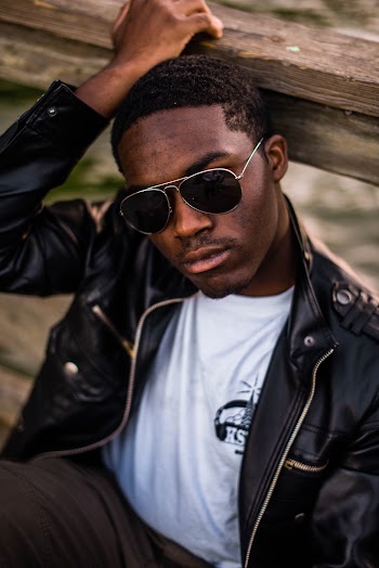 Photo of Man in Sunglasses and Black Leather Jacket Posing While Sitting By Wooden Railing