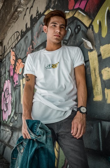 Photo of Man in White T-shirt and Gray Jeans Leaning on Graffiti Wall While Holding Denim Jacket