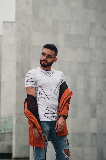 Photo of Man in White T-shirt, Orange Jacket, and Blue Jeans Posing In Front of Gray Wall