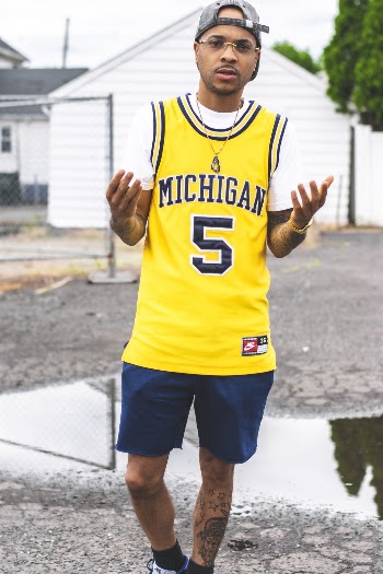 Photo of Man in Yellow Michigan Basketball Jersey, Blue Shorts, and Hat Standing on Concrete Pavement Posing With His Hands Out