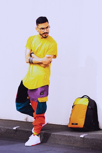 Photo of Man in Yellow T-shirt and Multicolored Pants Posing Beside Backpack Near White Wall
