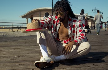 Photo of Man Wearing Red and Gray Plaid Dress Shirt and Beige Pants Holding Beige and Red Fedora Hat Sitting on Brown Wooden Boardwalk