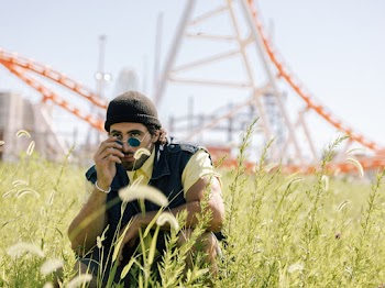 Photo of Man Wearing Yellow Crew-neck Shirt and Black Vest Crouching on Grass Field Holding Sunglasses