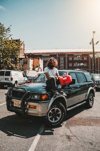 Photo of Man With His Her Over His Face Sitting on Hood of a SUV Parked in a Parking Lot Holding a Jerrycan