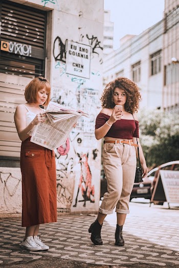 Photo of One Woman Reading a Newspaper and Another Holding Her Phone Standing by the Sidewalk
