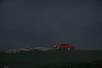 Photo of Red Sedan Parked  on a Grassy Field With Foggy Hills in the Background