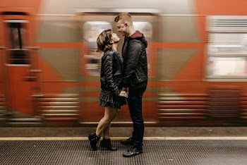 Photo of Smiling Couple Standing on Train Station Platform While Holding Hands as a Train Passes