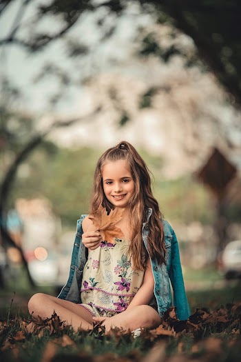 Photo of Smiling Girl Sitting on Ground Holding Out Dry Maple Leaf
