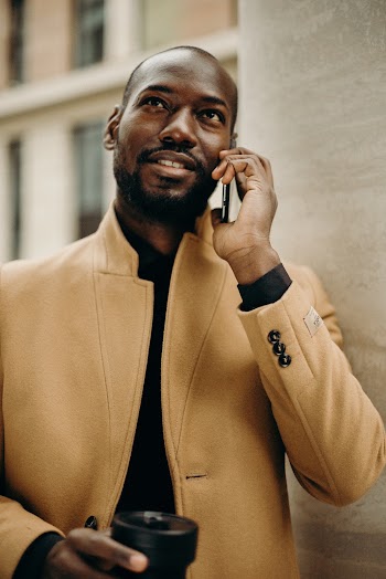 Photo of Smiling Man in Brown Coat Talking on the Phone and Holding a Cup While Standing Beside a White Column