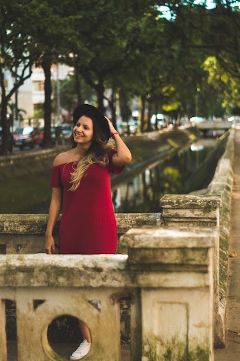 Photo of Smiling Woman in Black Hat and Red Dress Standing on Gray Concrete Bridge