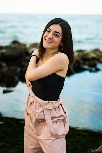 Photo of Smiling Woman in Black Top and Pink Dungarees Standing with Body of Water in the Background