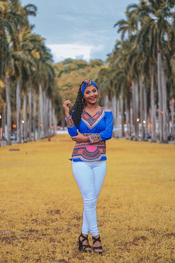 Photo of Smiling Woman in Blue Dashiki and White Jeans Standing on Yellow Grass Near Palm Trees Posing