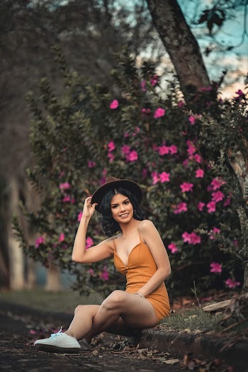 Photo of Smiling Woman in Mustard Deep V-neck Spaghetti-strap Dress  and Brown Fedora Hat Sitting on the Curb by Pink Flower Plant