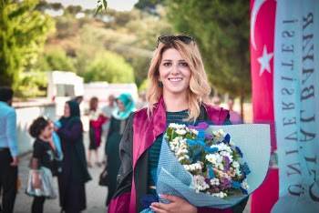 Photo of Smiling Woman in Pink and Black Academic Gown Posing While Holding Bouquet of Flowers with a Crowd of People in the Background