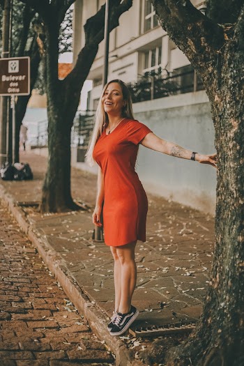 Photo of Smiling Woman in Red Dress Tiptoeing by Tree on Sidewalk