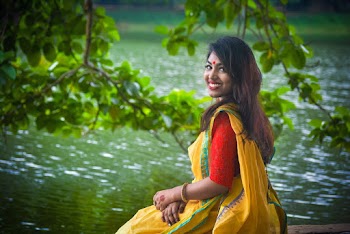 Photo of Smiling Woman in Red Lace Top and Yellow Saree Dress Sitting on Stone Surface by Body of Water